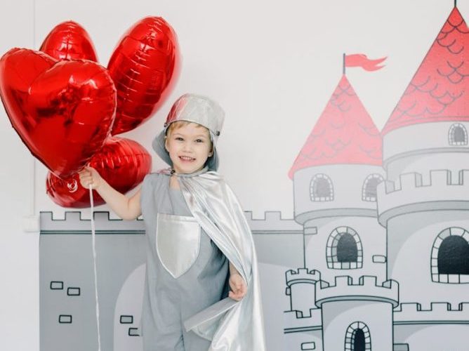 Ein fröhliches Kind im Ritterkostüm mit herzförmigen Luftballons steht drinnen vor einem Burgwandgemälde.